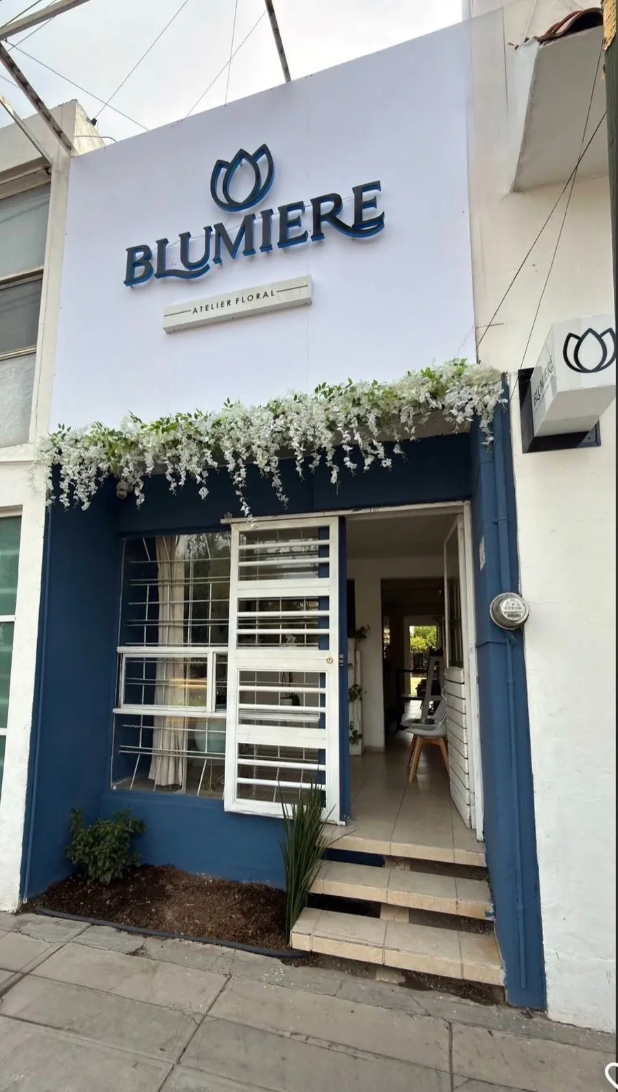 Fachada azul del atelier Blumiere, con el letrero y flores blancas sobre la entrada
